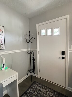 A room with a white door, a coat rack, a small table with a clock, and a patterned rug. - Renovated Brick Cottage · 10 Min to Uptown (Charlotte)