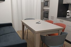 A compact kitchen with a microwave, oven, and coffee machine, a dining table with two chairs, and a sofa in the foreground.