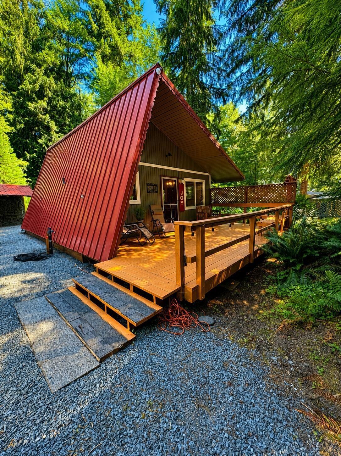 A small cabin with a red roof, a wooden deck, and a gravel driveway.