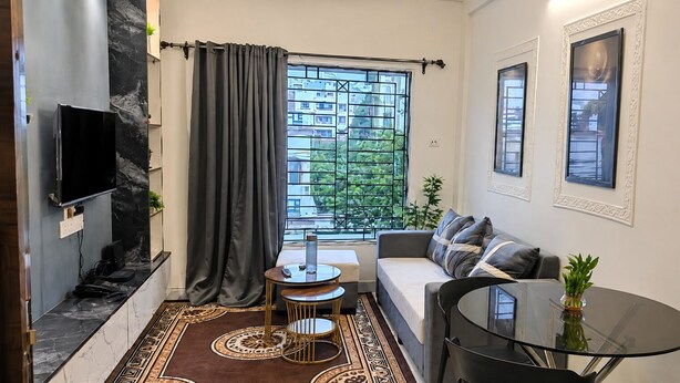 A compact living space with a sofa, a small round table, a TV mounted on the wall, and a window with a view of greenery.
