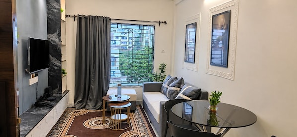 A compact living space with a sofa, a small round table, a TV mounted on the wall, and a window with a view of greenery.