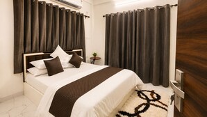A hotel room with a bed, curtains, an air conditioning unit, a small table with a plant, and a wooden door.