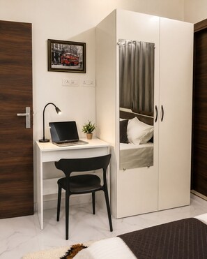 A bedroom with a bed, a desk with a laptop, a chair, a wardrobe, and a potted plant.