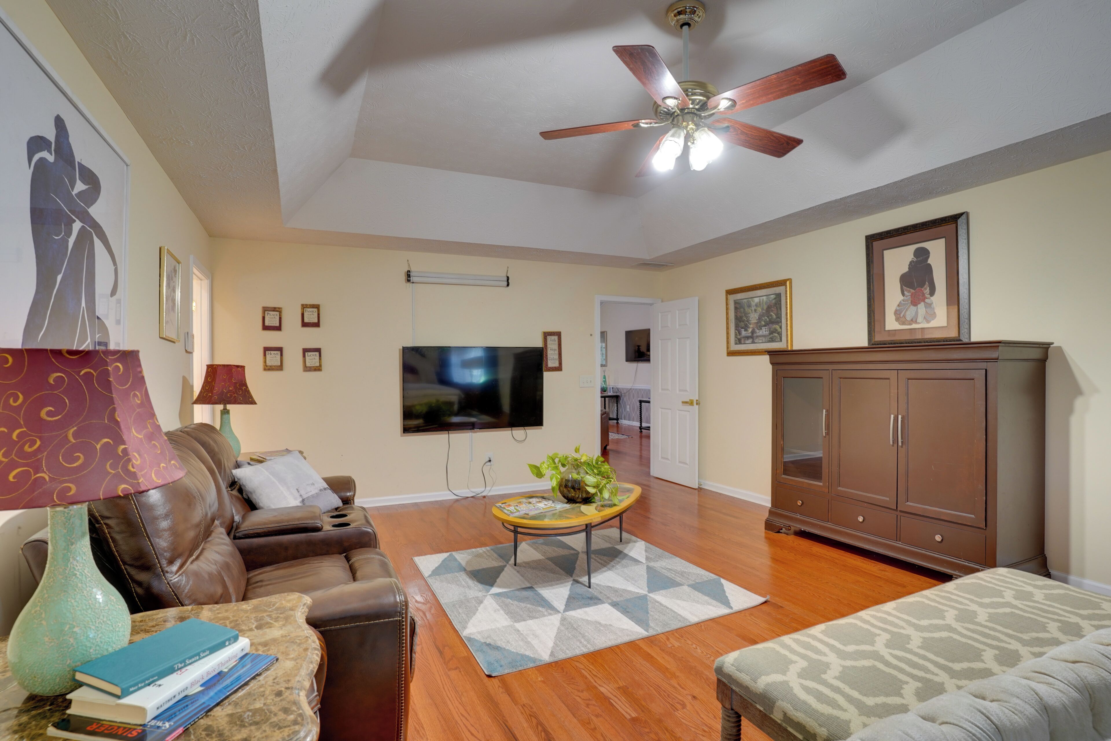A living room with a ceiling fan, a flat-screen TV, a brown leather sofa, a coffee table with books, and a patterned rug.