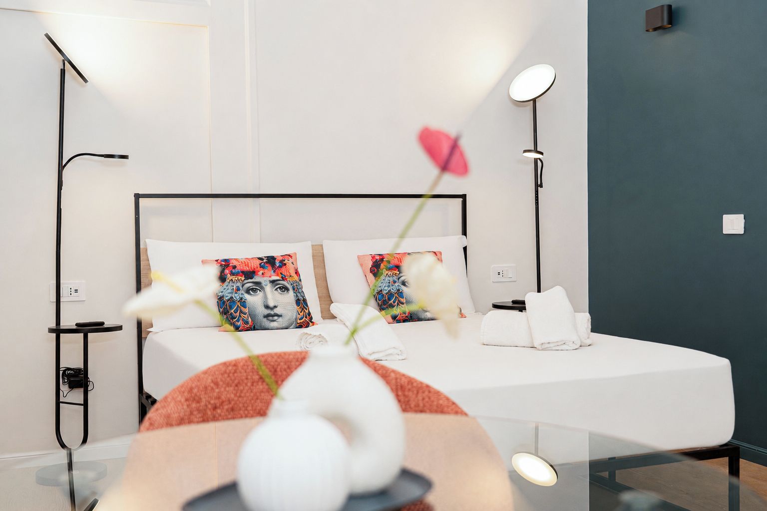 Una habitación de hotel moderna con cama, lámparas de noche, un florero con flores y un aplique de pared.