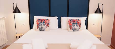 Una cama bien hecha con dos almohadas, dos lámparas de noche y un par de toallas en el suelo.