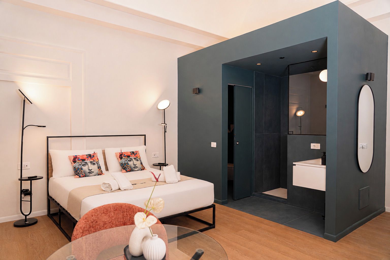 A modern hotel room with a bed, bedside lamps, a round table with a vase, and a view into the bathroom.