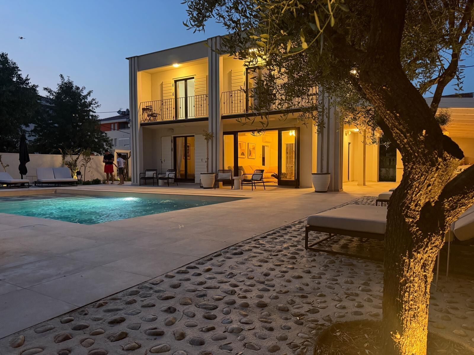 Una casa a due piani con piscina, area salotto esterna e un albero sullo sfondo.