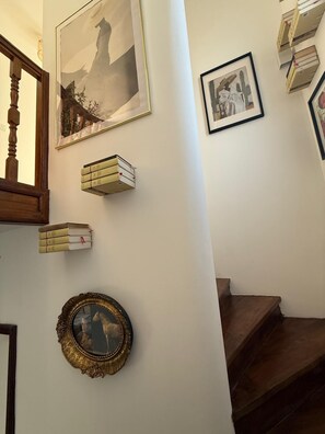 Un escalier avec une rampe et des marches en bois, un tableau encadré représentant un paysage, un portrait encadré et une pile de livres sur le mur.