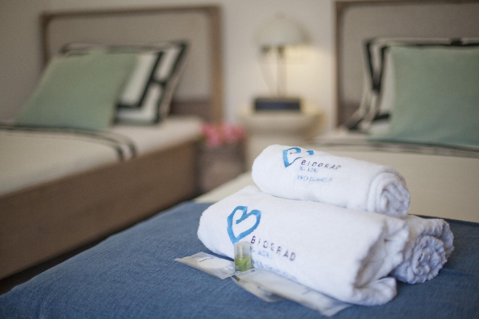 Ein Hotelzimmer mit zwei Betten, jeweils mit Kissen und einem Nachttisch mit Lampe. Auf dem Bett liegen Handtücher mit einem Herz-Logo.