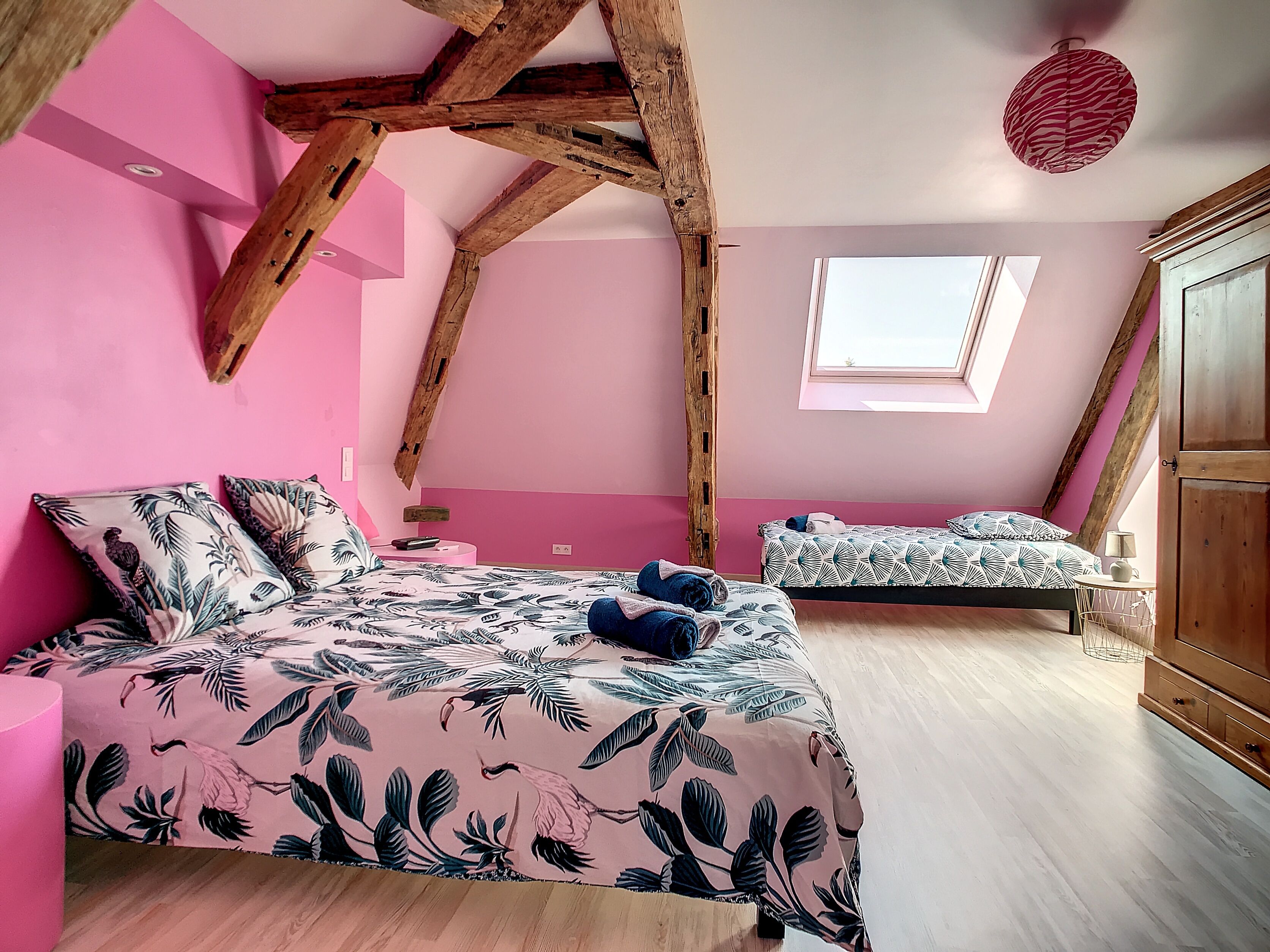 A bedroom with a sloped ceiling, a bed with a floral bedspread, a smaller bed, a window, and a wooden wardrobe.