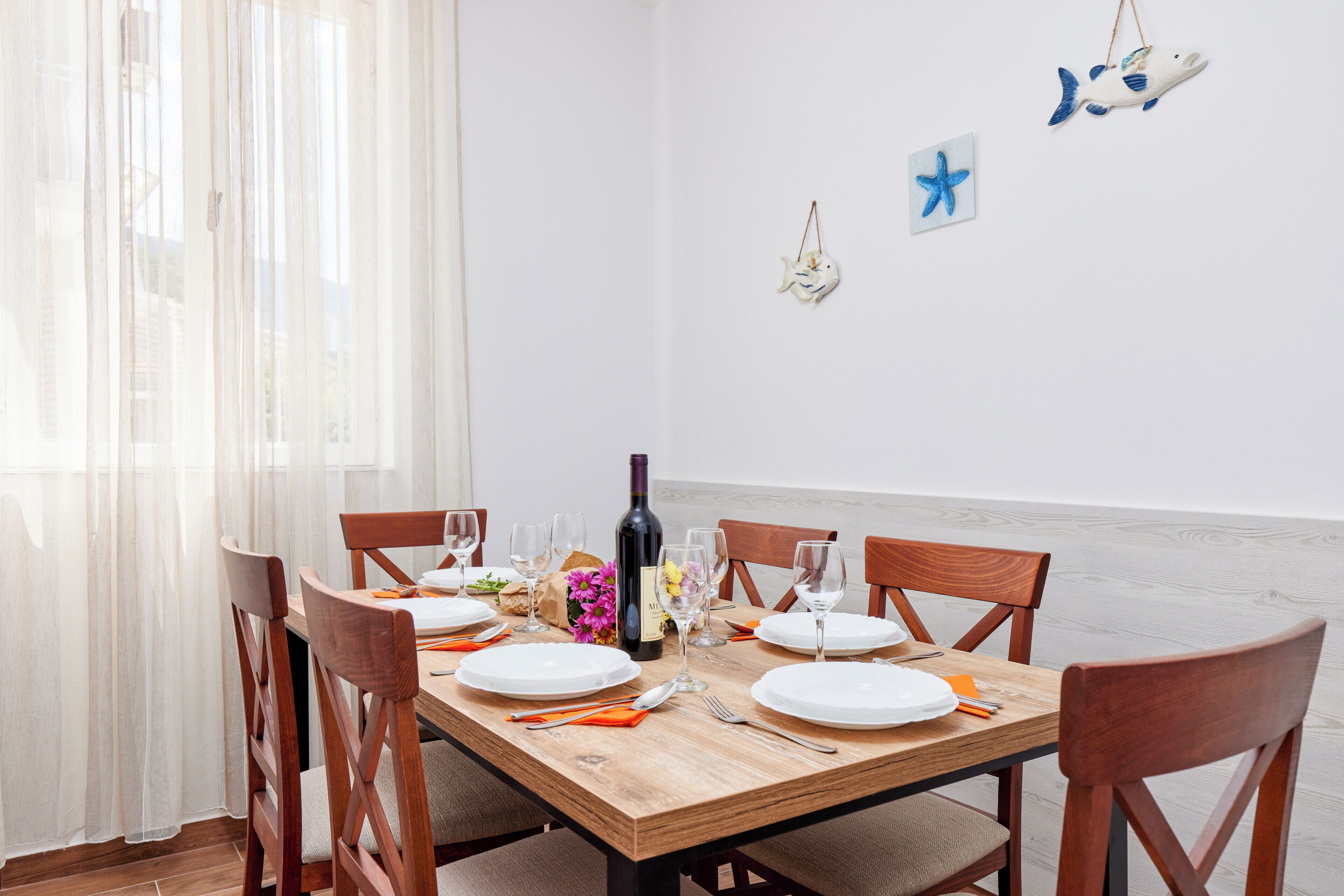 Mesa de comedor preparada para seis personas con sillas de madera, una botella de vino y un florero con flores.
