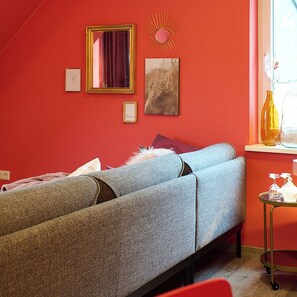 A room with a bed, a window with curtains, a small table with a vase, and wall decorations including a mirror and framed pictures. - Just like home, but in a completely different place (Bengel)