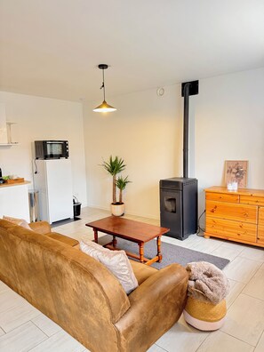 Ein Wohnzimmer mit einer Couch, einem Holztisch, einer Topfpflanze, einem Kamin und einer Küchenzeile mit Mikrowelle und Kühlschrank.