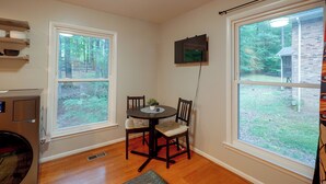 A room with a dining table and chairs, two windows, a wall-mounted TV, and a radiator. - Fire Pit & Grill! Wooded Durham Retreat (Durham)