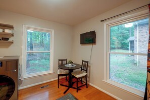 A room with a dining table and chairs, two windows, a wall-mounted TV, and a radiator. - Fire Pit & Grill! Wooded Durham Retreat (Durham)