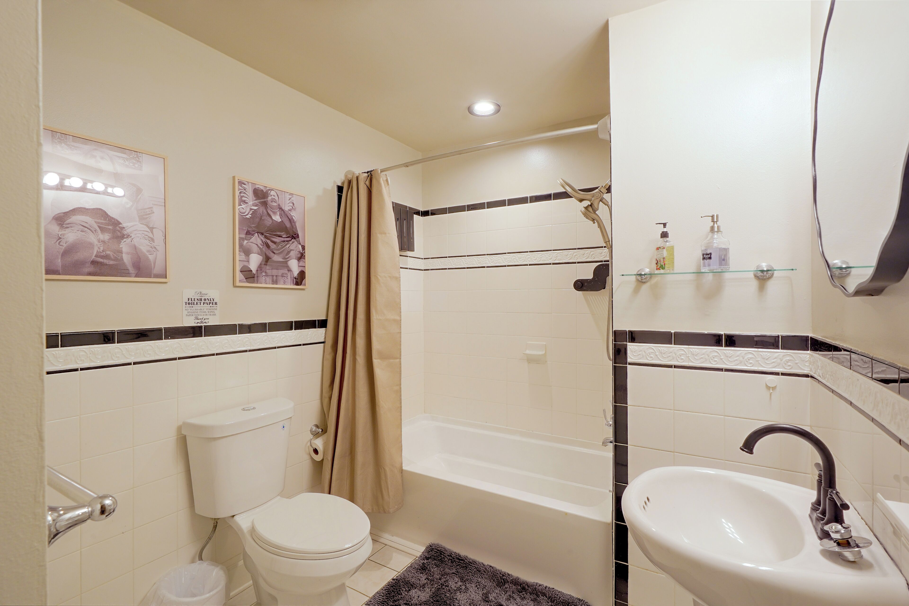 A bathroom with a toilet, bathtub, and sink. There are two framed pictures on the wall, a shower curtain, and a glass shelf with toiletries.