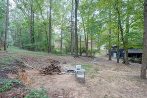 A forested backyard with a fire pit, a chair, and a stack of firewood. - Fire Pit & Grill! Wooded Durham Retreat (Durham)