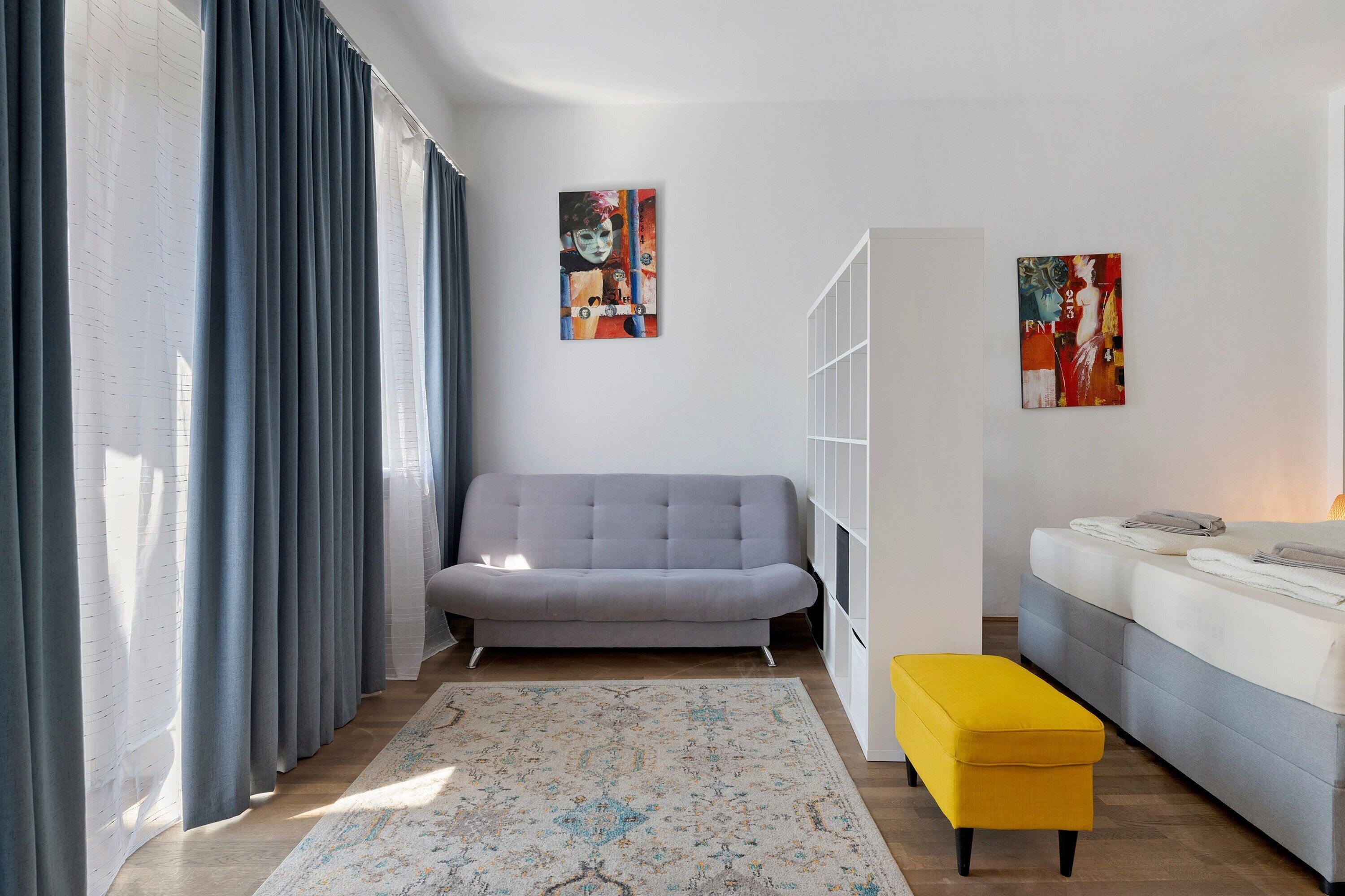 Ein modernes Wohnzimmer mit einem grauen Sofa, einem gelben Hocker und zwei Bildern an der Wand.