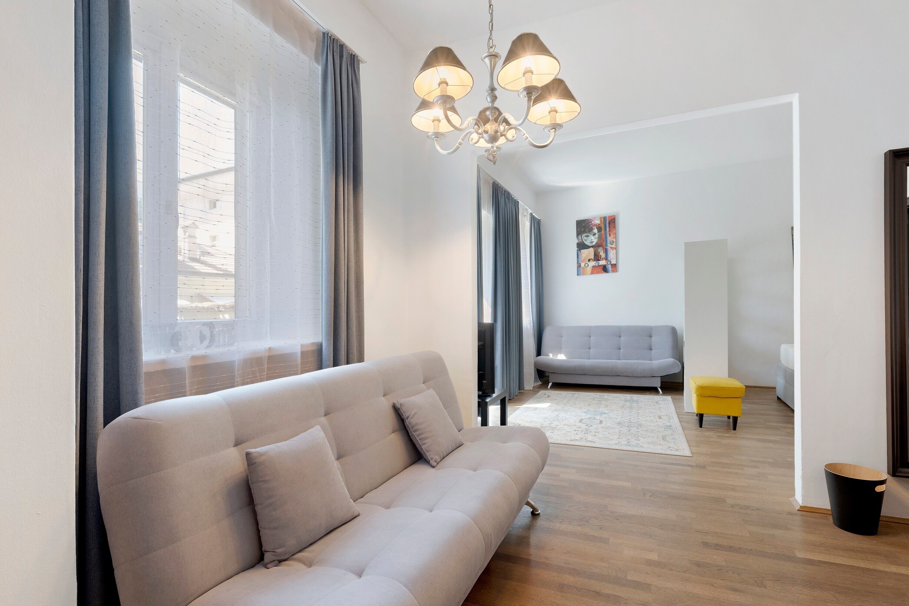 A modern living room with a sofa, a chandelier, a grey sofa, a painting, and a yellow ottoman.