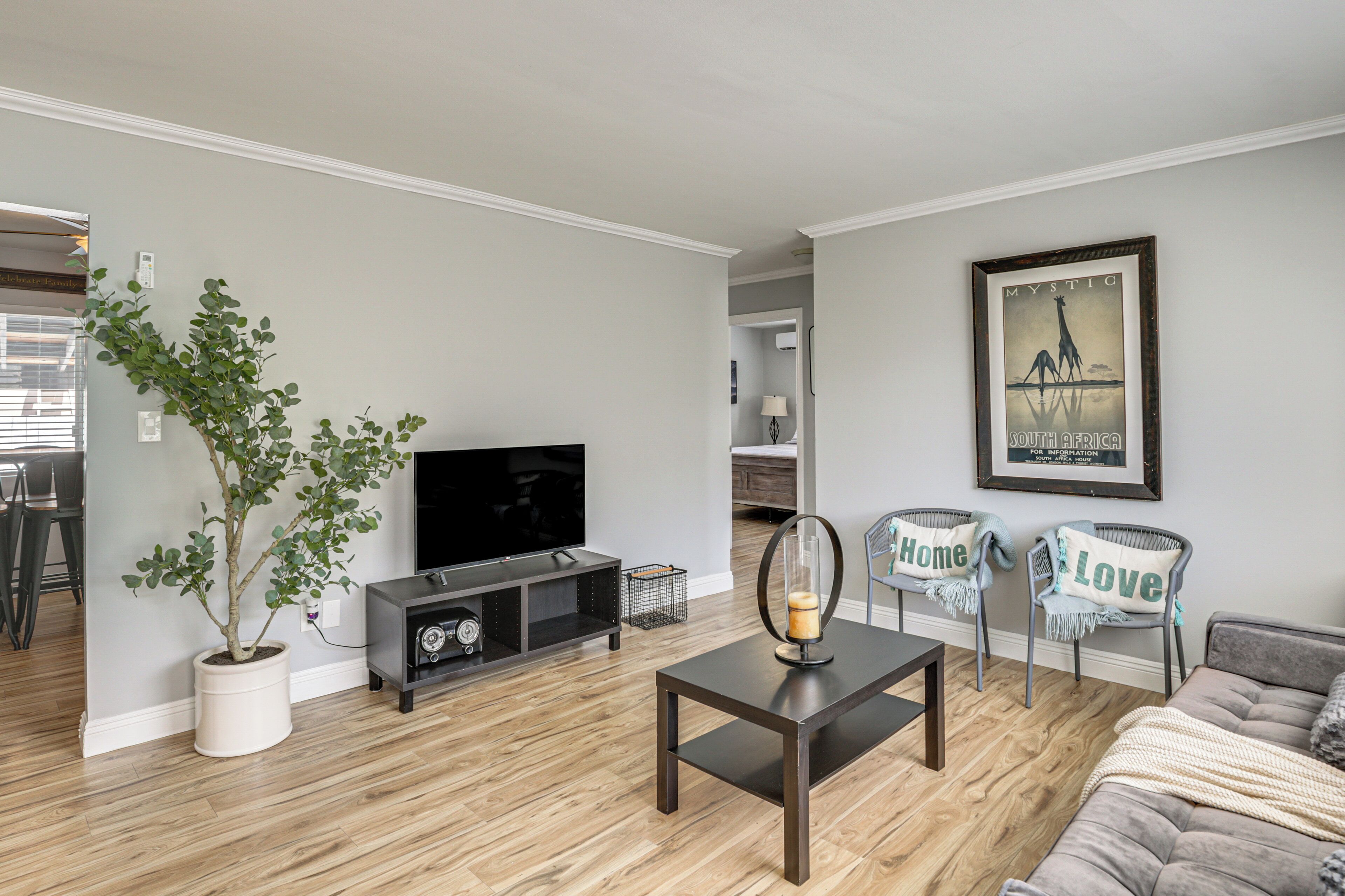 A modern living room with a flat-screen TV, a coffee table, and a sofa. There is a potted plant and a framed picture on the wall.