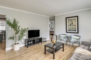 A modern living room with a flat-screen TV, a coffee table, and a sofa. There is a potted plant and a framed picture on the wall.