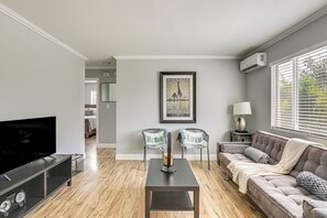 Ein modernes Wohnzimmer mit Fernseher, Couchtisch, Sofa und zwei Sesseln. An der Wand hängt ein gerahmtes Bild und über dem Sofa befindet sich eine Klimaanlage.