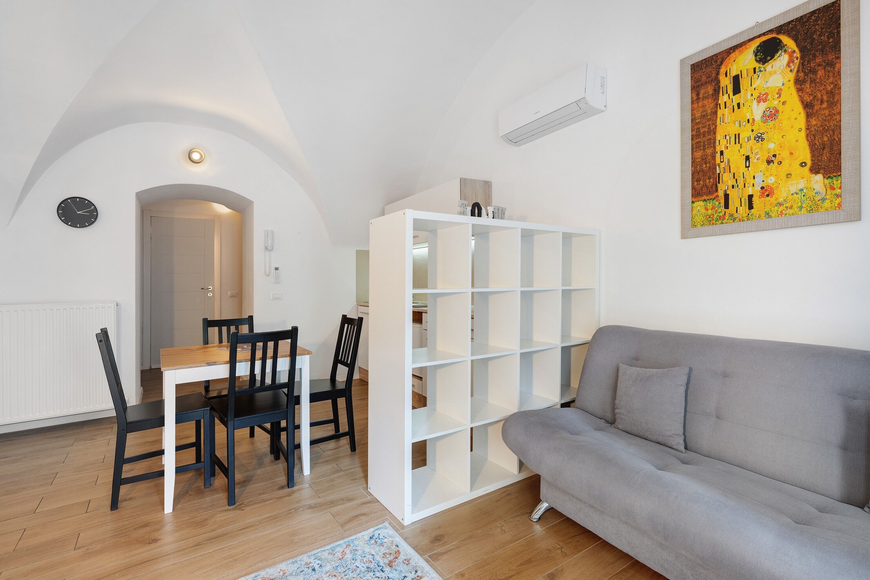 Uma sala de estar moderna com um sofá cinzento, uma mesa de jantar com cadeiras, uma estante branca e um quadro na parede.