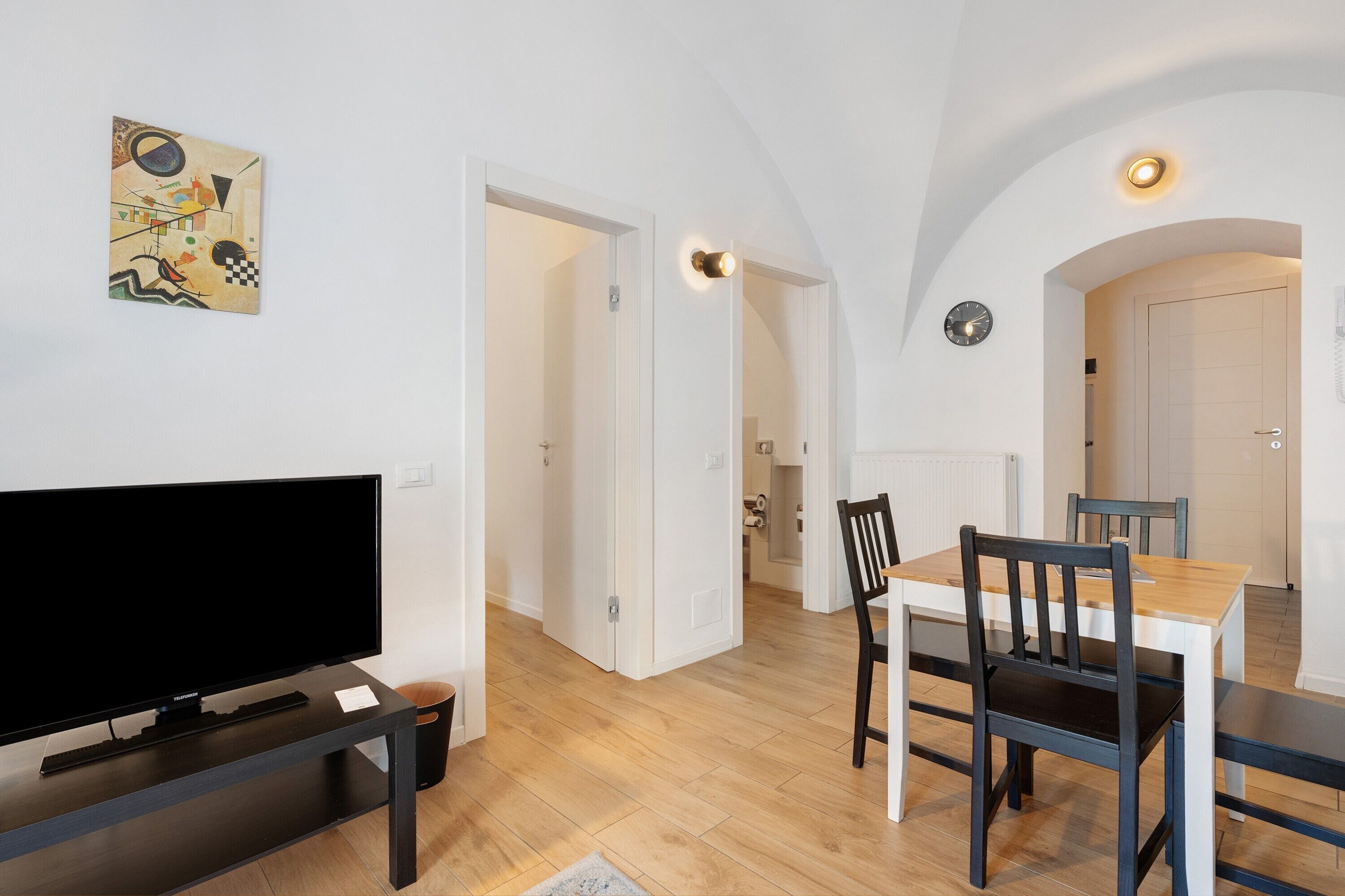 Een moderne woonkamer met een flatscreen-tv, een eetgedeelte met een houten tafel en stoelen, en een keuken die door een open deur zichtbaar is.
