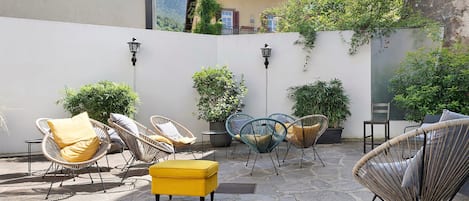 Een terras met rieten stoelen en kussens, planten in potten en een vloer van natuursteen.