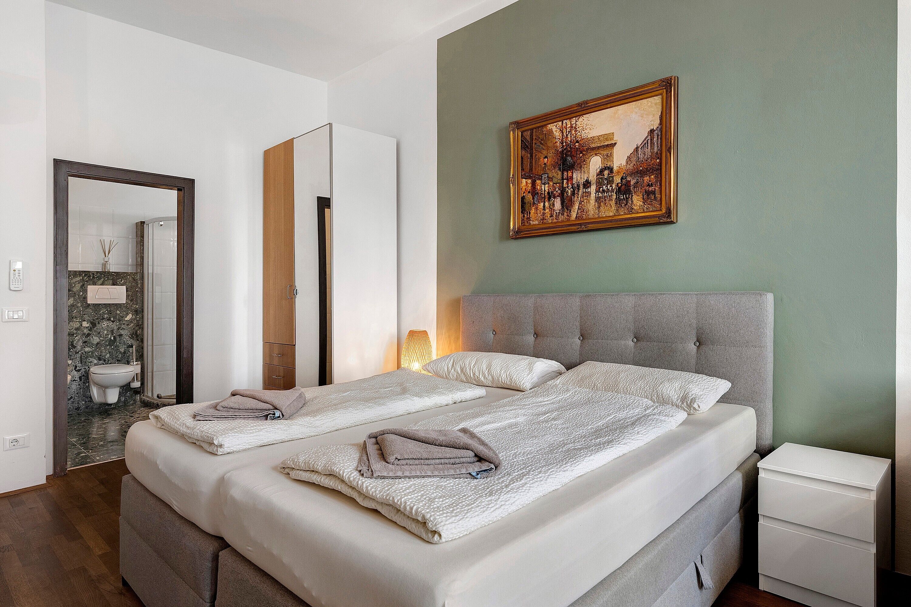 Un lit double avec une tête de lit grise, une table de chevet, un tableau accroché au mur et une salle de bain visible à travers une porte ouverte.