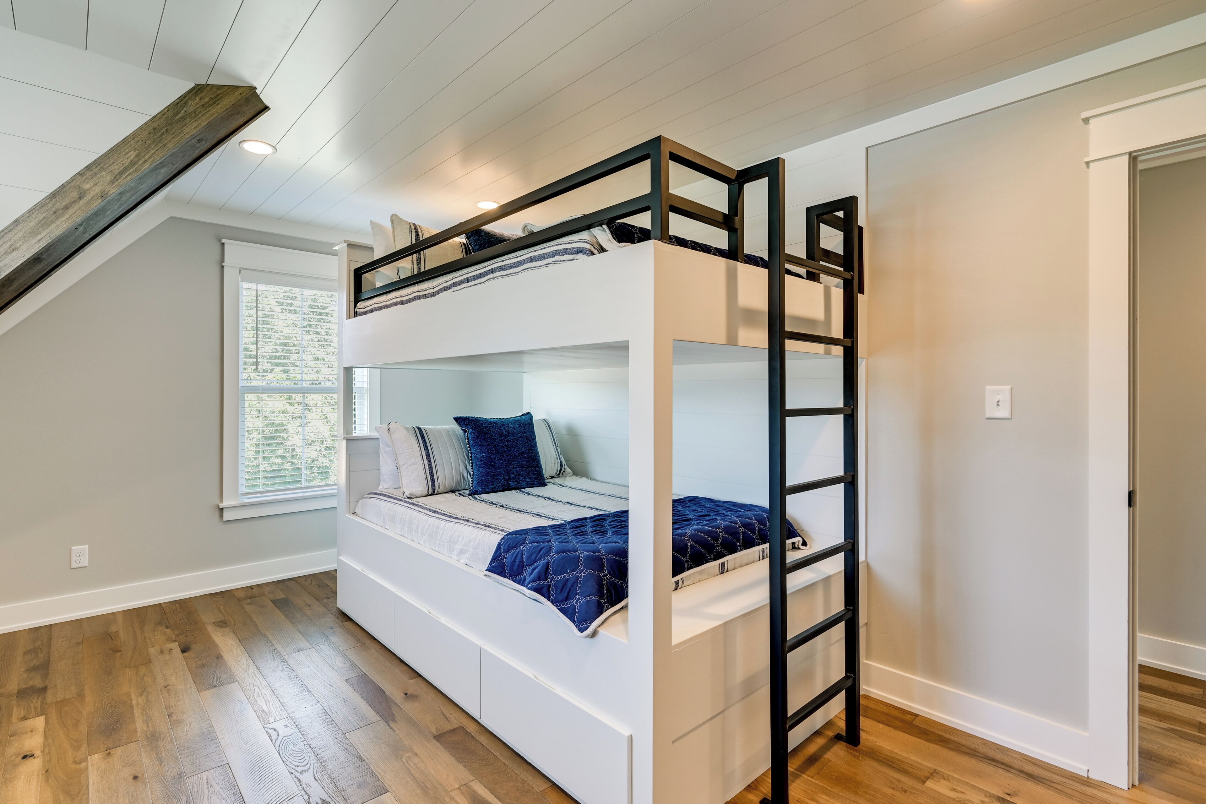 Un lit superposé avec une échelle, une fenêtre avec des stores et un plancher en bois.