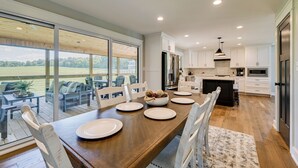 A modern kitchen with white cabinets, a dining area with a wooden table, and a view of a green landscape through large windows. - The Lily Pad - Nolin Lake Retreat (Clarkson)