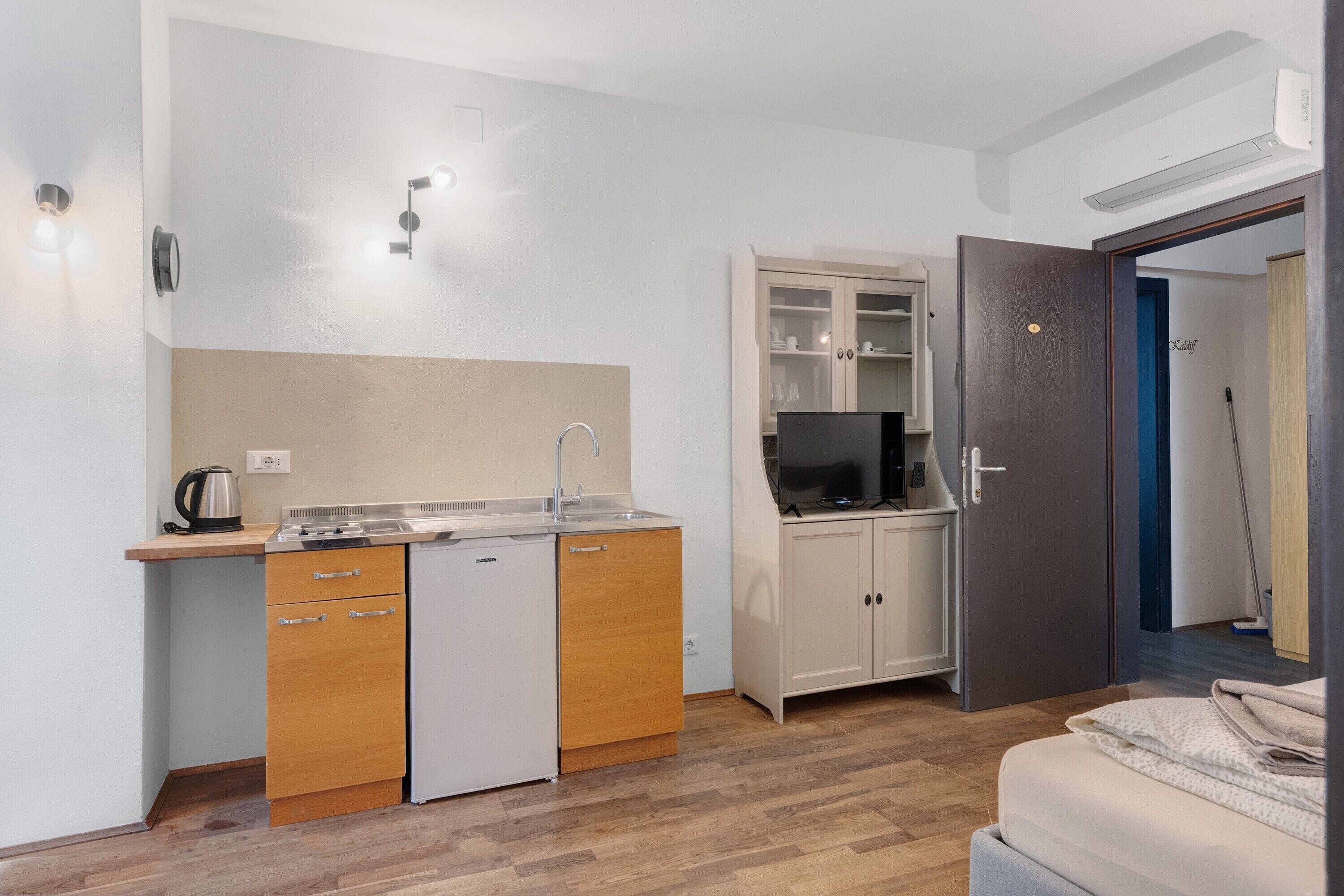 A compact kitchen with a sink, a small refrigerator, and a microwave. There's a cabinet with glass doors and a television on top.