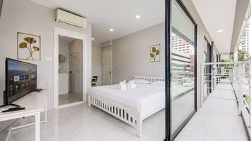 A modern hotel room with a bed, a desk with a computer, a bathroom, and a balcony with a city view.