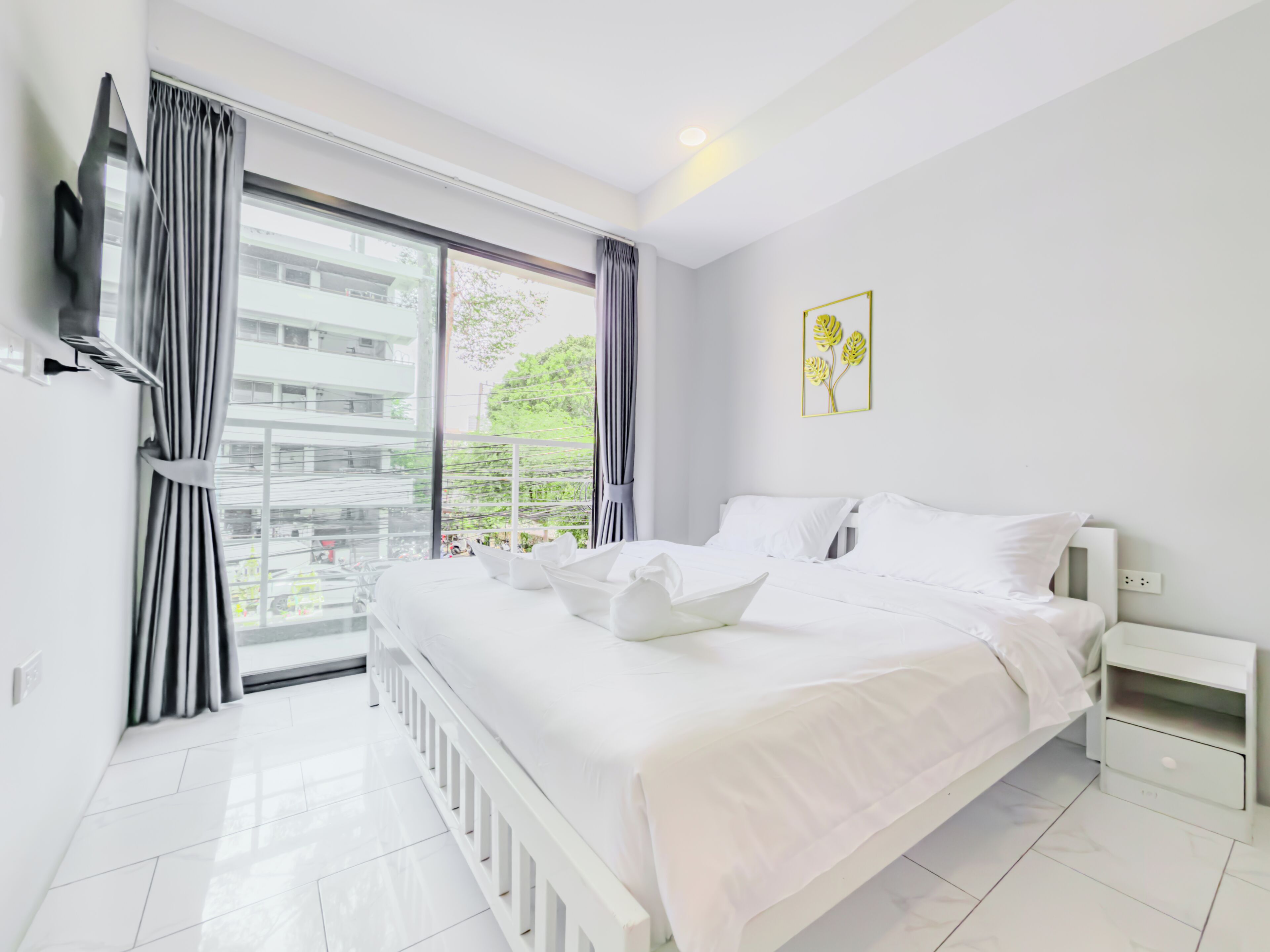 Una habitación de hotel moderna con una cama grande, una mesita de noche, un televisor montado en la pared y un ventanal con cortinas.