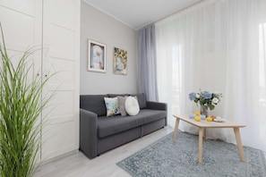 A modern living room with a grey sofa, two framed artworks, a small table with flowers, and a large window with sheer curtains. - Stylish Studio with a Balcony Central Warsaw (Warszawa)