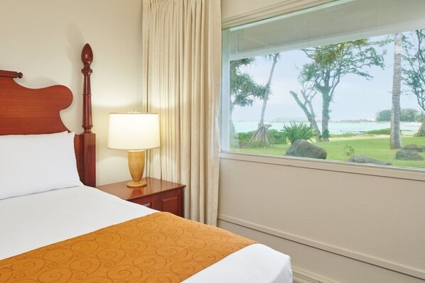 Ein Hotelzimmer mit Bett, Nachttisch mit Lampe, Fenster mit Blick auf Bäume und Strand.