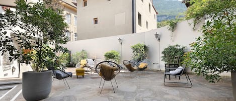 An outdoor seating area with stone flooring and wicker chairs, surrounded by greenery and a building with a tiled roof.