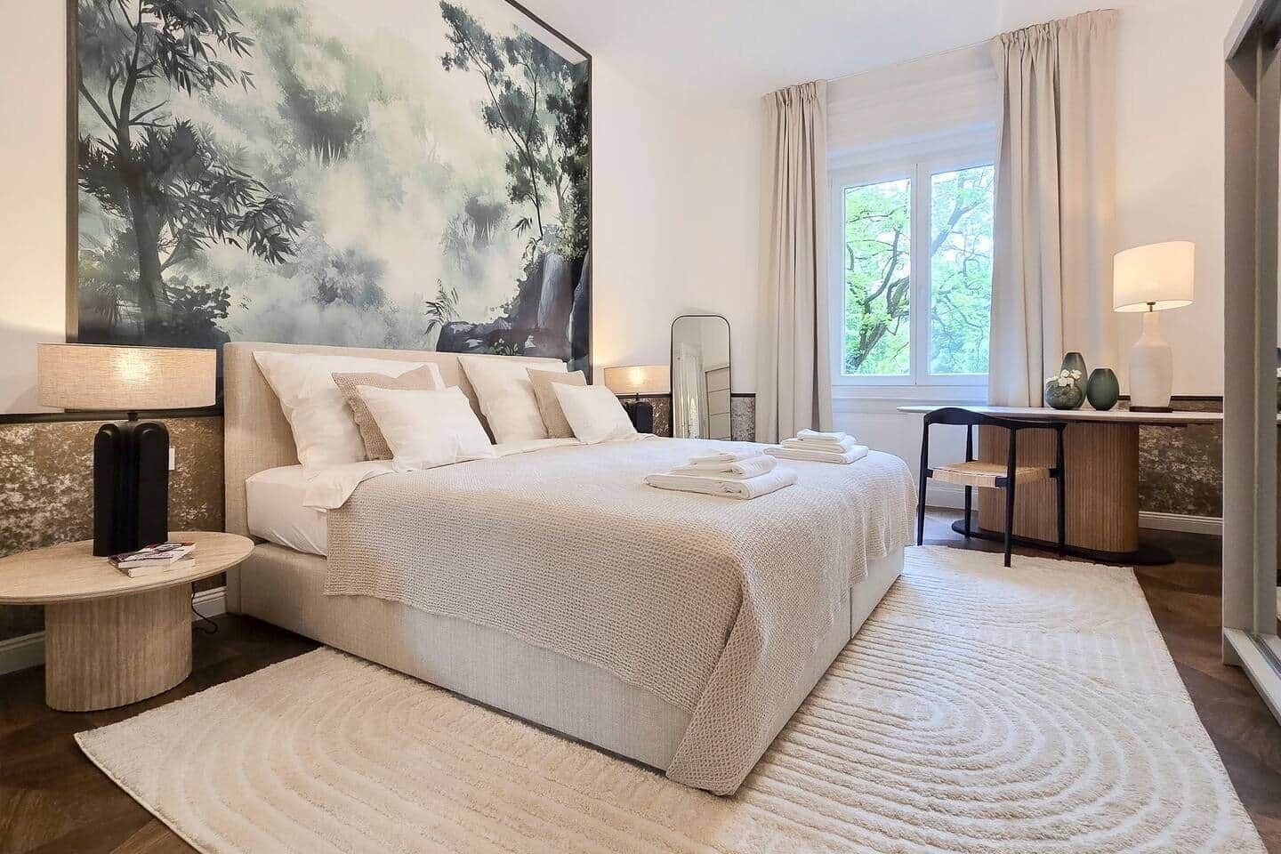 A bedroom with a large bed, a bedside table with a lamp, a nightstand, a window with curtains, and a painting on the wall.
