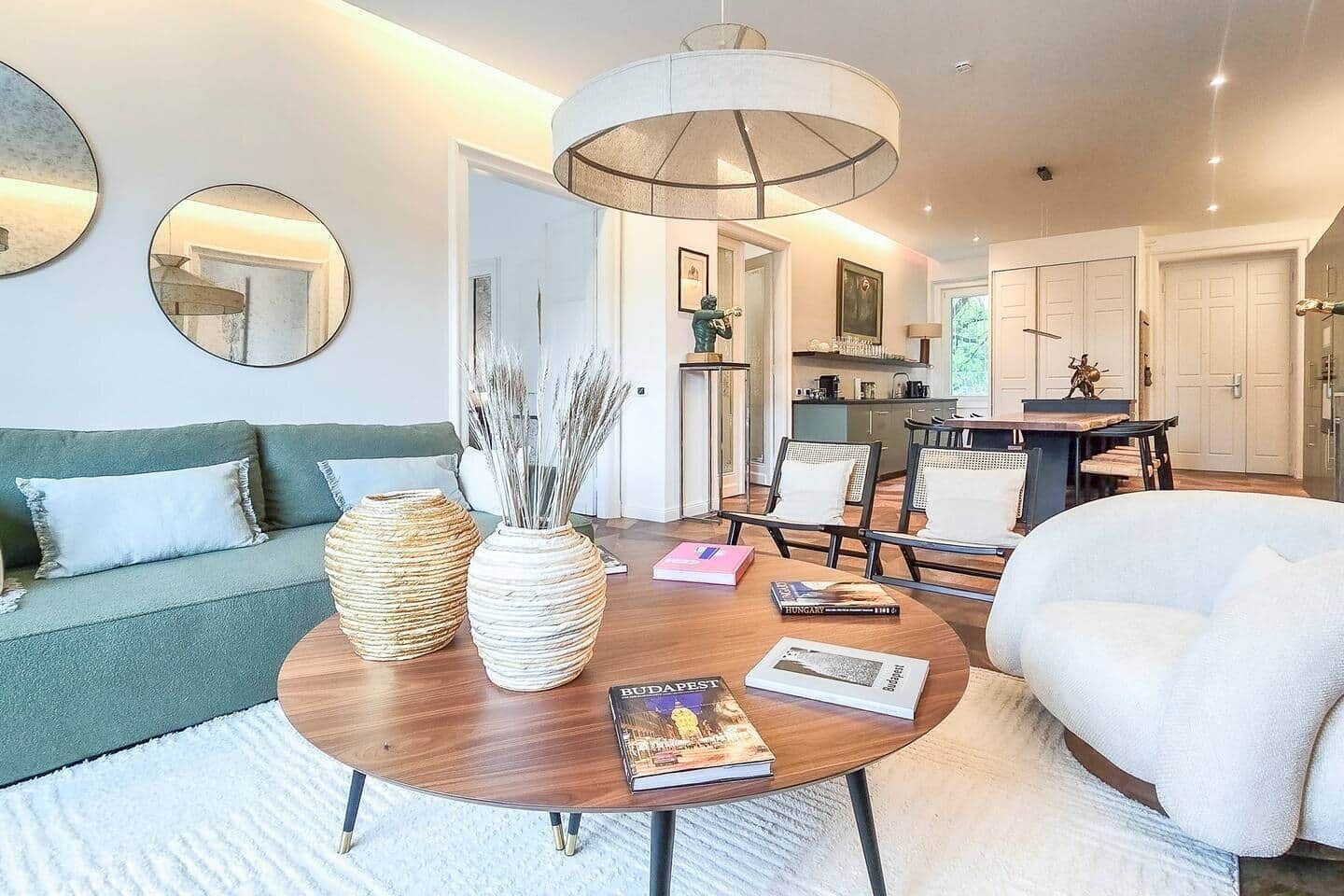 A modern living room with a round wooden coffee table, a sofa, and a white armchair. There are two large vases on the table, and a book titled 'HUNGARY' is visible.
