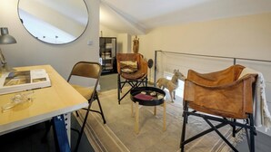 Um quarto com um espelho redondo, uma mesa de madeira com uma lâmpada, duas poltronas de couro, uma mesa redonda pequena e uma lousa.
