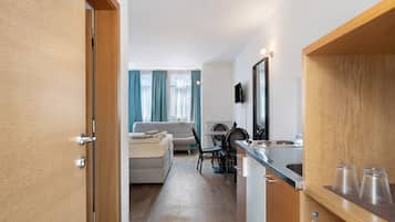 Habitació d'hotel moderna amb una porta de fusta, una cuineta amb taulell d'acer inoxidable, zona de dinar amb taula i cadires, i zona de estar amb un sofà i una televisió.