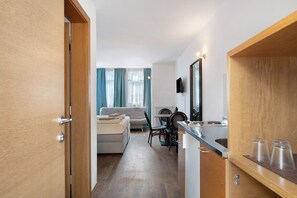 Habitació d'hotel moderna amb una porta de fusta, una cuineta amb taulell d'acer inoxidable, zona de dinar amb taula i cadires, i zona de estar amb un sofà i una televisió.
