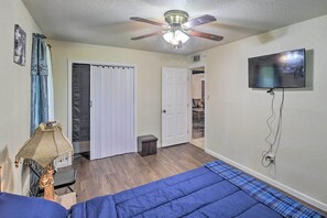 Een slaapkamer met een plafondventilator, een bed met blauw beddengoed, een aan de muur bevestigde televisie en een deur die naar een andere kamer leidt.