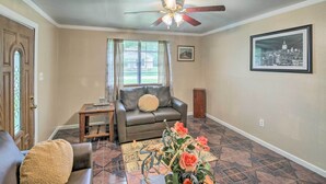A living room with a brown leather couch, a coffee table with flowers, a ceiling fan, and a window with curtains. - Family Home + Guest House! Near Fishing, Boat Ramp (West Orange)