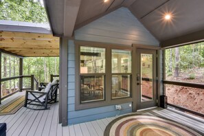 A covered deck with a seating area, large windows, and a view of the surrounding forest. - Tiny Home - Fire Pit, Grill, Deck & View! (Franklin)