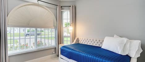 A bedroom with a bed, a ceiling fan, a window with blinds, and a view of a parking lot.