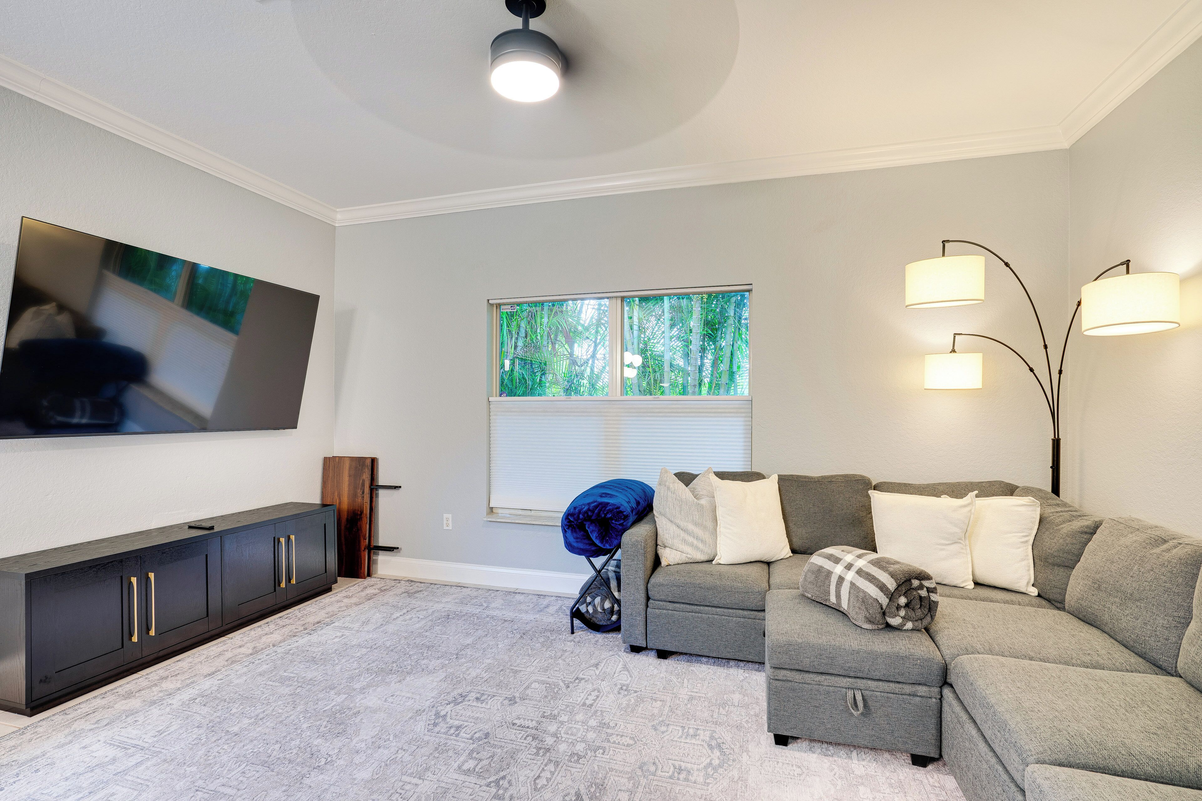 A living room with a grey sofa, a flat-screen TV, a dark wooden cabinet, and a window with blinds.