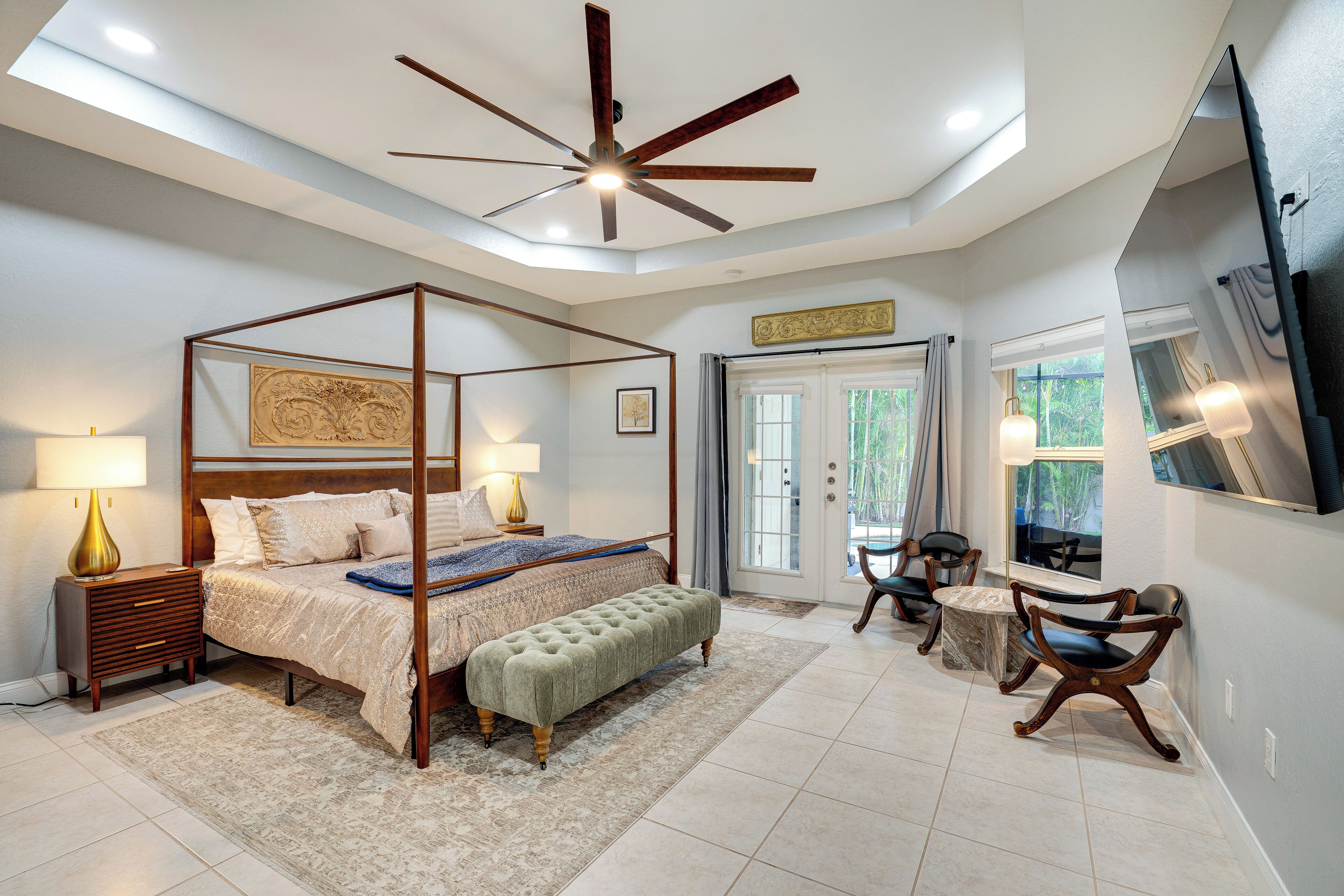 A bedroom with a four-poster bed, a sitting area with chairs, a television, and a ceiling fan.